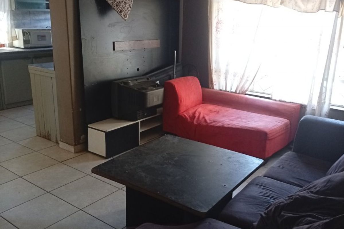 A living room area featuring tiled flooring and a built-in wall panel, with an adjacent view into the kitchen space.