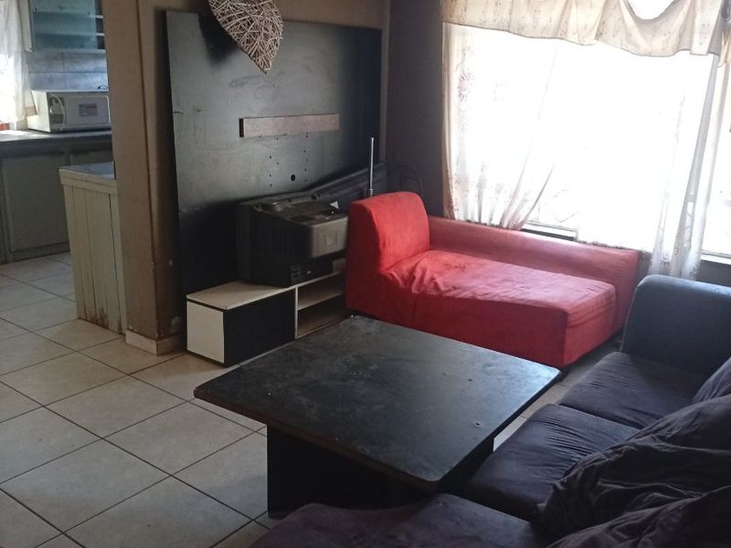 A living room area featuring tiled flooring and a built-in wall panel, with an adjacent view into the kitchen space.