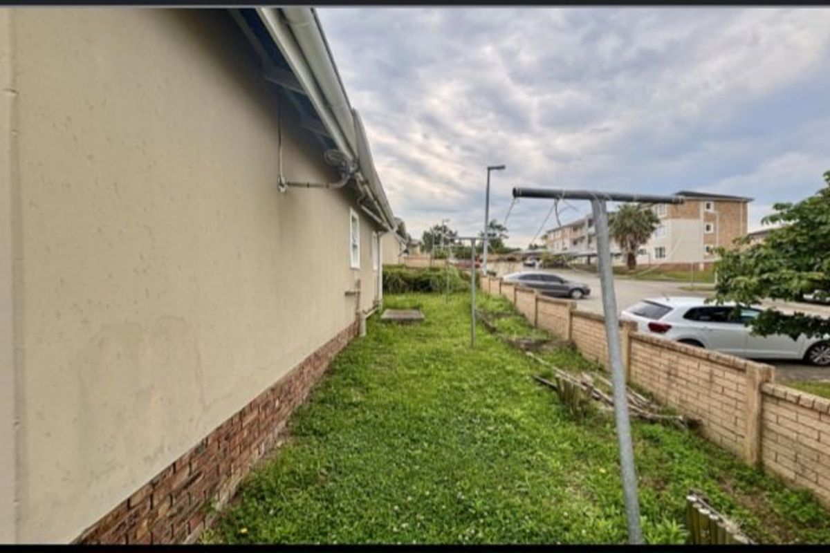 A side exterior view of the townhouse featuring a plastered wall with brick plinth, adjacent to a grassed area and brick boundary wall.