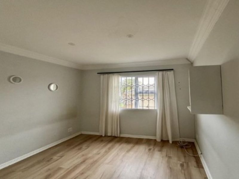 A bright, empty bedroom featuring light wood-look laminate flooring, cove cornicing, and a window fitted with security bars.