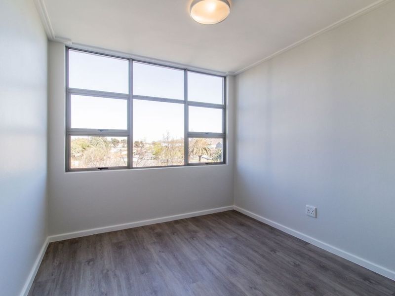 A bright, empty bedroom featuring modern laminate flooring, a large window with natural light, and a neutral color palette.
