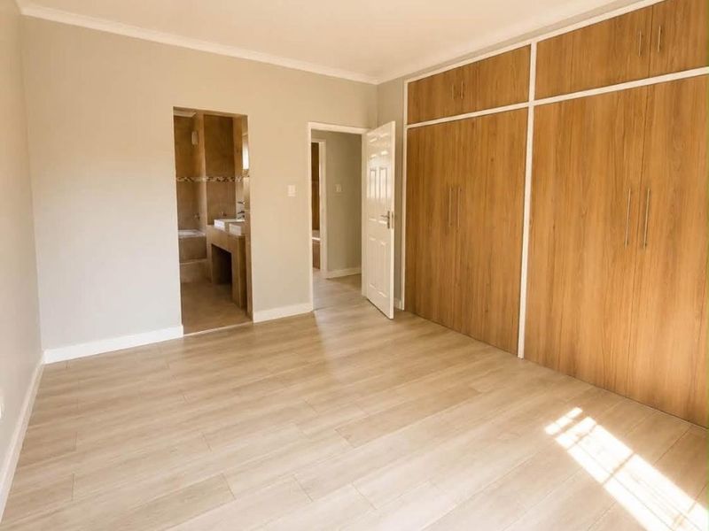 A bright, empty bedroom featuring modern laminate flooring, extensive built-in wooden cupboards, and direct access to an en-suite bathroom.