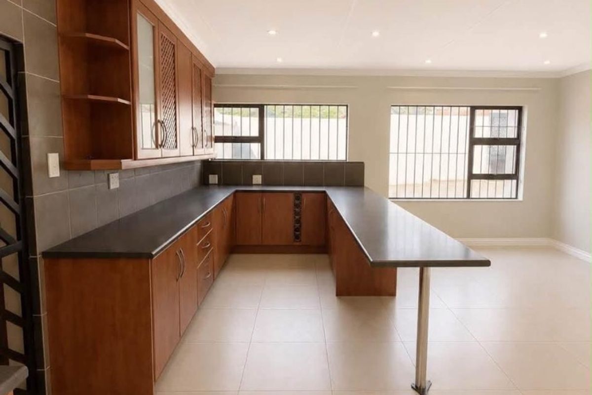 A spacious, modern kitchen featuring warm-toned wooden cabinetry, dark stone countertops, a breakfast bar, and neutral floor tiles in a clean, open-plan layout.
