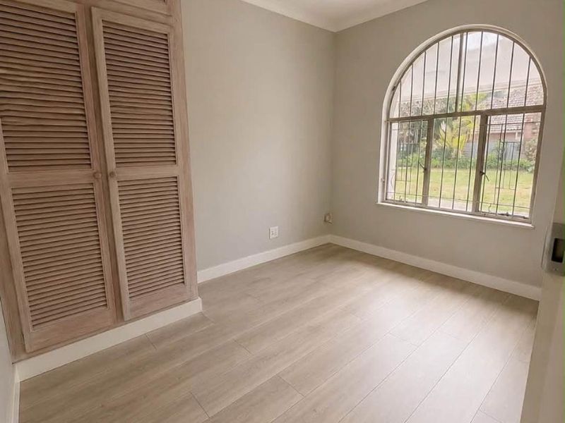 A bright, empty bedroom featuring light laminate flooring, a built-in louvered wardrobe, and a distinctive arched window with security bars.