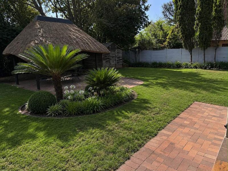 A well-maintained garden featuring a traditional thatched lapa, brick paved pathways, and established landscaped garden beds with a lush lawn.
