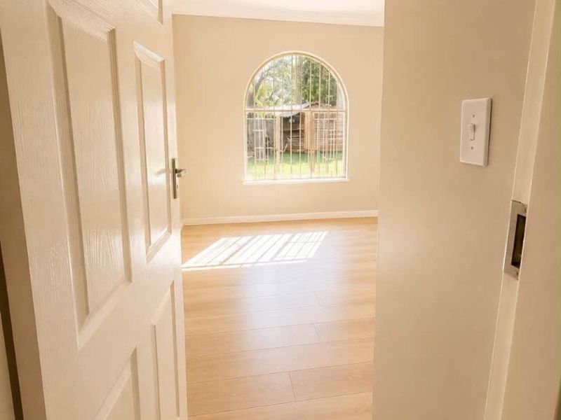 A bright, empty bedroom featuring light-toned laminate flooring, an arched window with security bars, and neutral wall finishes.