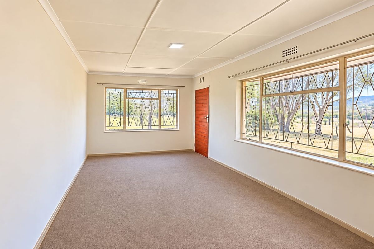 A bright, empty living room featuring neutral carpeting, large windows with security bars, and a wooden door, offering a clean canvas for personalization.