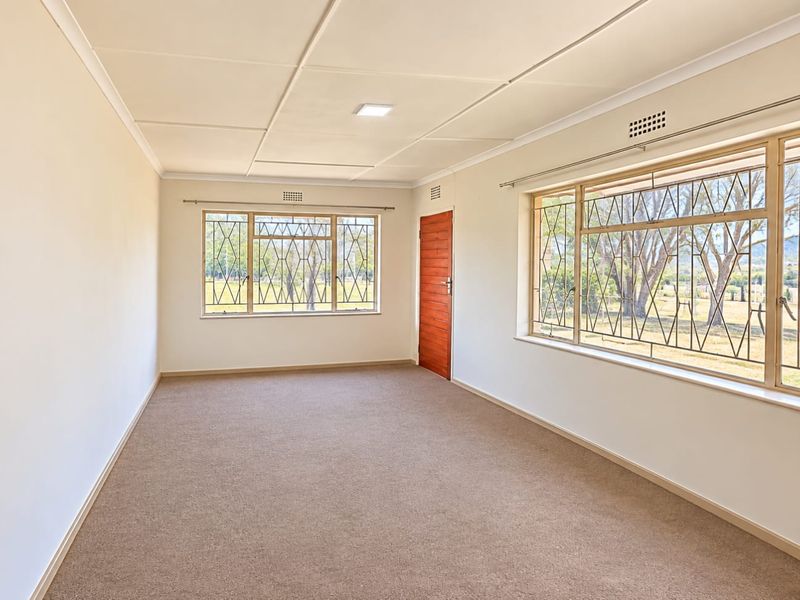 A bright, empty living room featuring neutral carpeting, large windows with security bars, and a wooden door, offering a clean canvas for personalization.