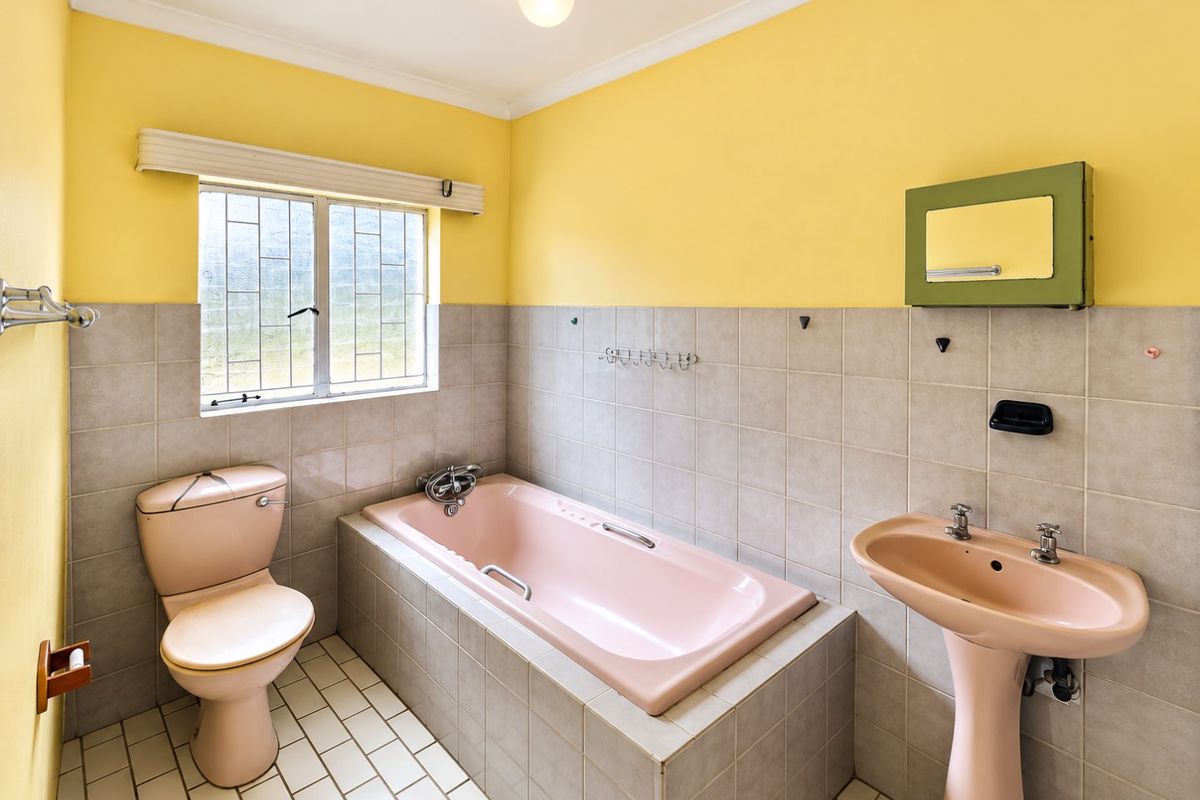 A dated bathroom featuring a pink pedestal basin, matching bathtub, and toilet, complemented by neutral wall tiles and yellow painted upper walls.