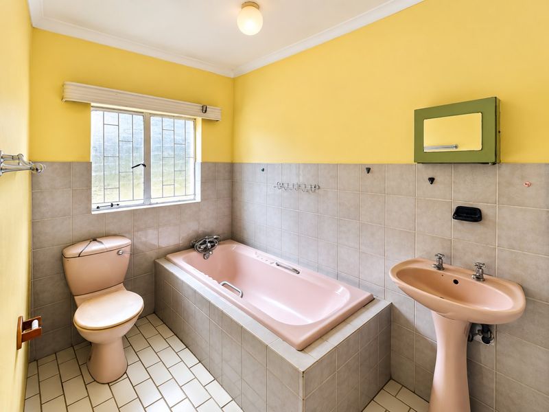 A dated bathroom featuring a pink pedestal basin, matching bathtub, and toilet, complemented by neutral wall tiles and yellow painted upper walls.