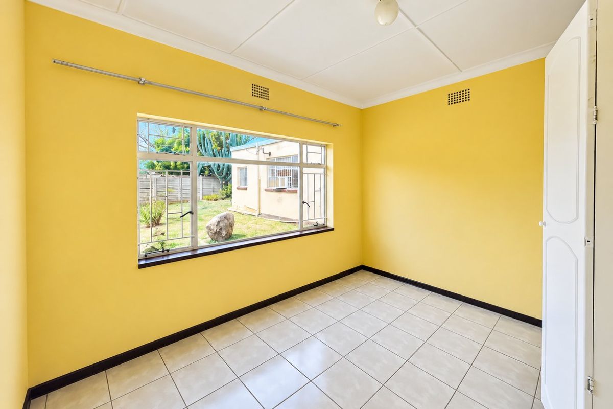 An empty bedroom featuring bright yellow walls, tiled flooring, and a large window with security bars overlooking the rear garden.