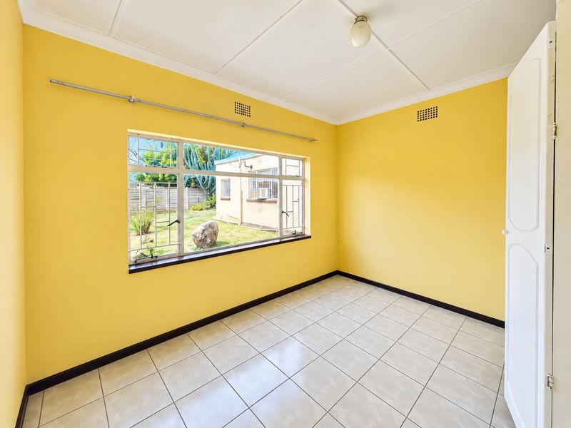 An empty bedroom featuring bright yellow walls, tiled flooring, and a large window with security bars overlooking the rear garden.