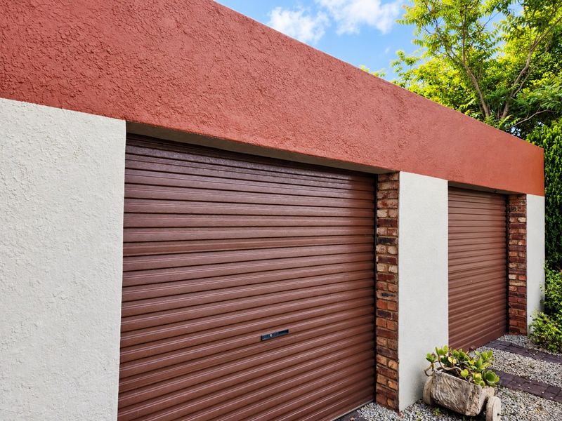 A well-maintained double garage featuring brown roller shutter doors, exposed brick detailing, and a contrasting red and white stucco exterior finish.