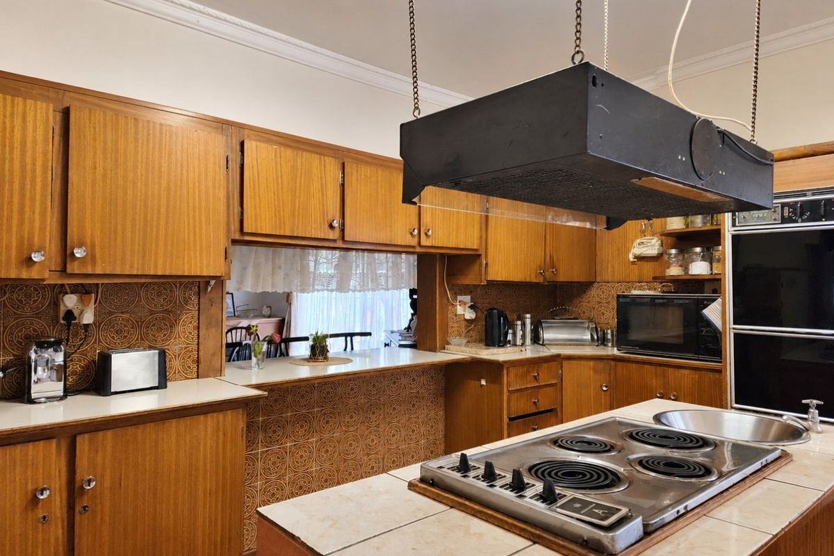 A vintage-style kitchen featuring extensive wooden cabinetry, a central island with an electric cooktop, and decorative tiled backsplashes, offering significant potential for modern renovation.