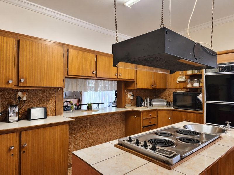 A vintage-style kitchen featuring extensive wooden cabinetry, a central island with an electric cooktop, and decorative tiled backsplashes, offering significant potential for modern renovation.