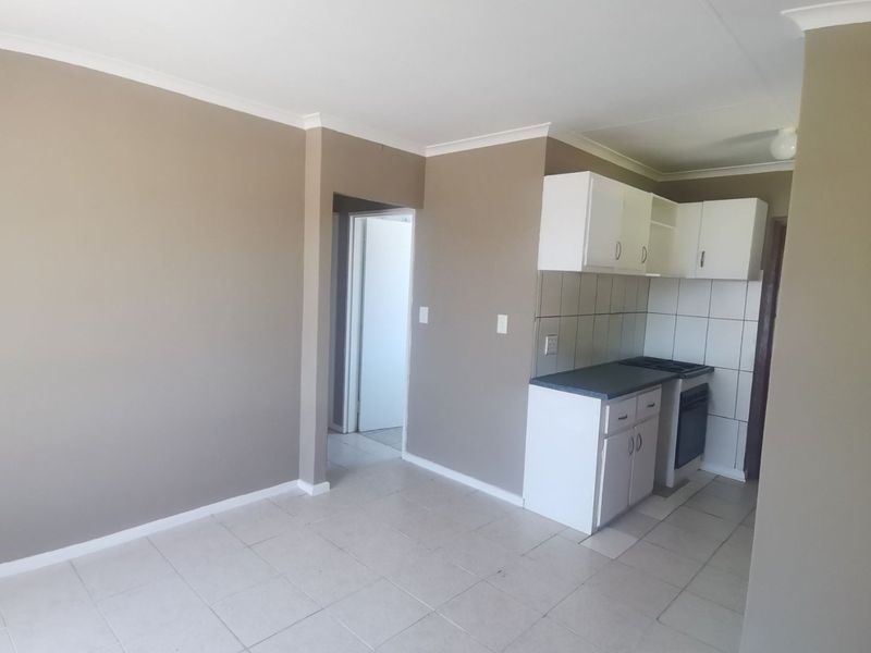 An open-plan living area featuring a compact kitchen with built-in cabinetry, oven, and tiled flooring, leading to an adjacent room.