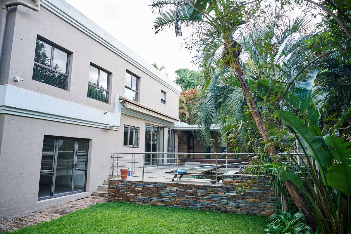 Rear view of the property featuring a paved patio with a stone-clad retaining wall, stainless steel balustrade, and established garden surroundings.
