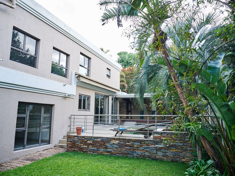 Rear view of the property featuring a paved patio with a stone-clad retaining wall, stainless steel balustrade, and established garden surroundings.