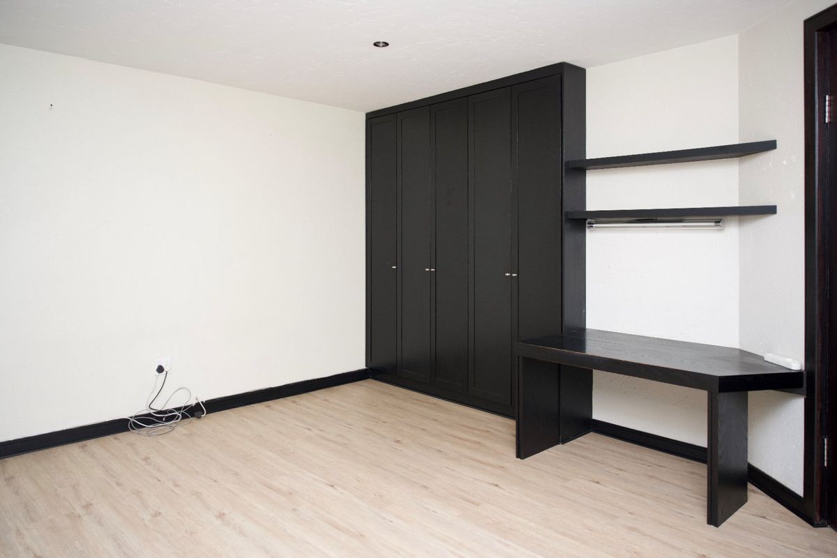 A clean, empty bedroom featuring light wood-look laminate flooring, a large black built-in wardrobe, and a matching built-in desk with floating shelves.
