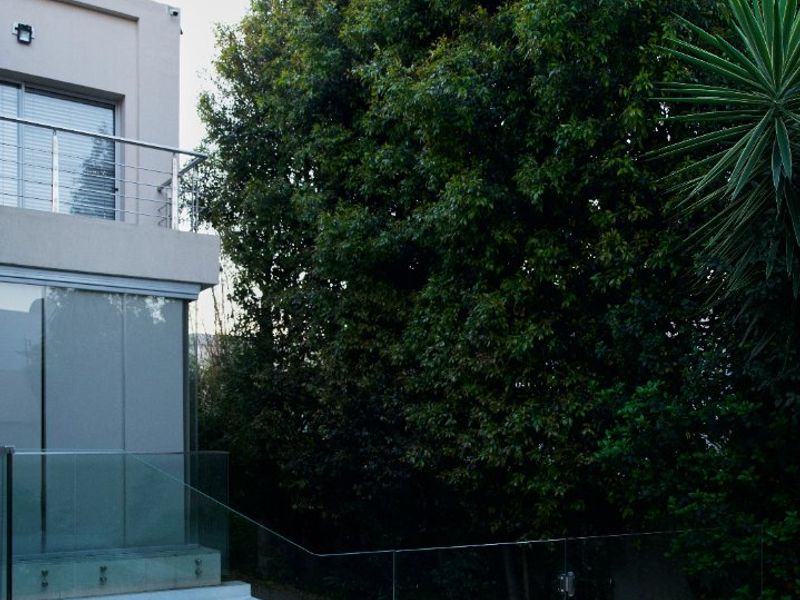 A modern swimming pool area featuring a glass balustrade, tiled surround, and an adjacent balcony, set against a backdrop of mature trees.
