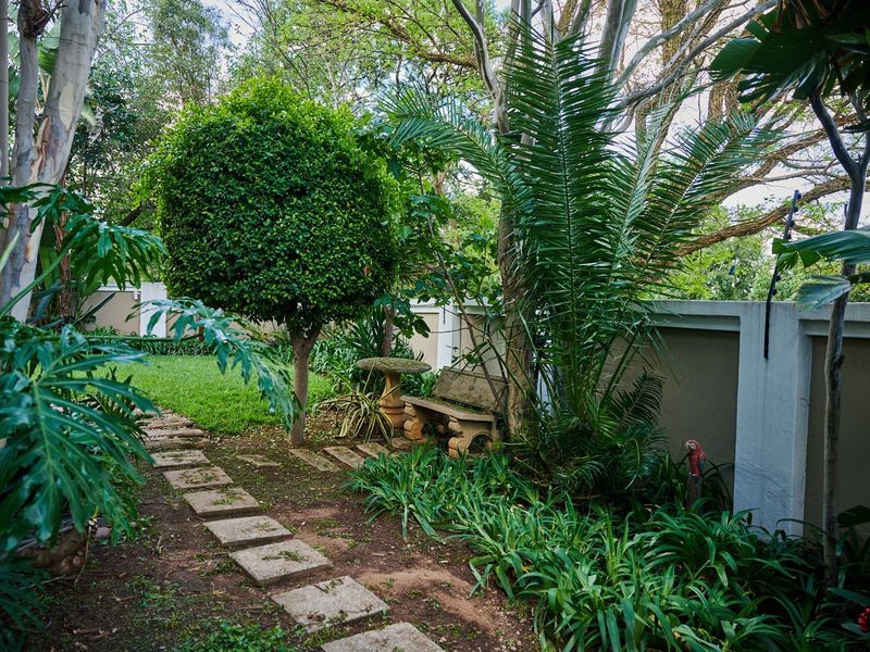A lush, well-established garden featuring a stone paver pathway, mature trees, and a secure perimeter boundary wall.