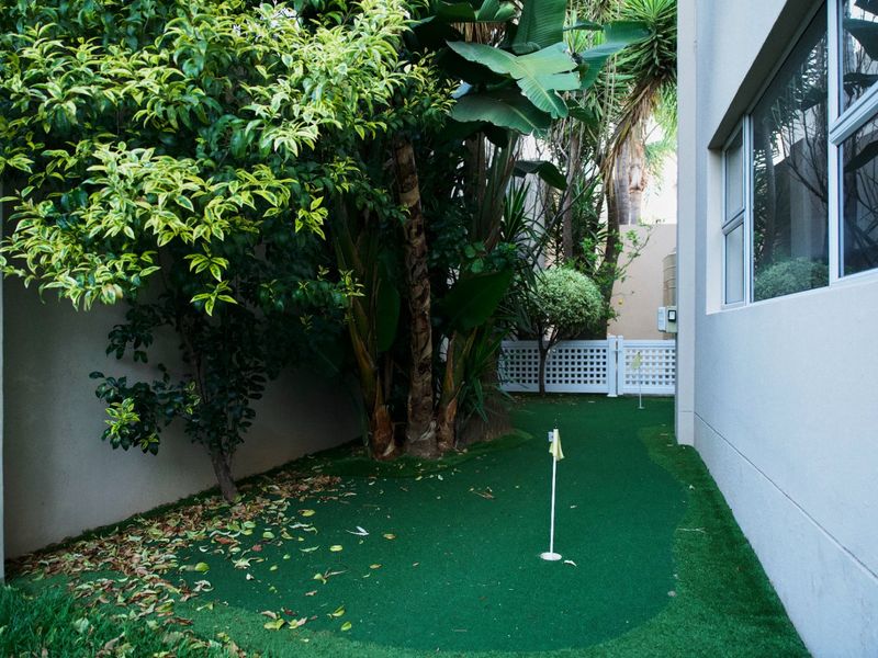 A private side garden area featuring a compact artificial turf putting green with two holes, bordered by established trees and a white lattice fence.