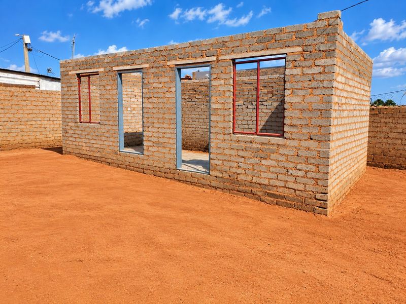 An unfinished brick structure showing the exterior shell with installed steel window and door frames, set on a cleared plot of land.