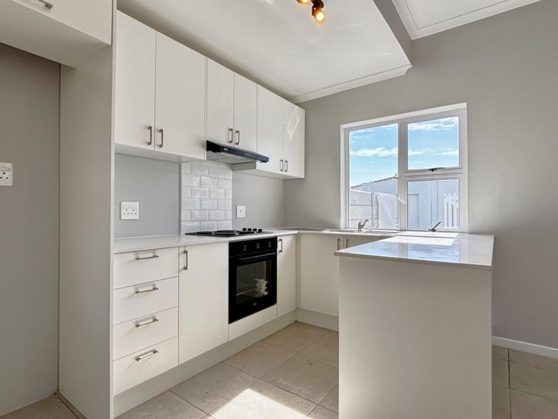 A modern, clean kitchen featuring white cabinetry, stone countertops, built-in oven and hob, subway tile backsplash, and neutral tiled flooring.