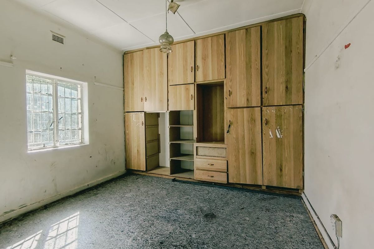 A vacant bedroom featuring built-in wooden cupboards, security bars on the window, and worn carpeting, requiring cosmetic updates and refurbishment.