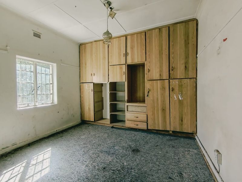 A vacant bedroom featuring built-in wooden cupboards, security bars on the window, and worn carpeting, requiring cosmetic updates and refurbishment.