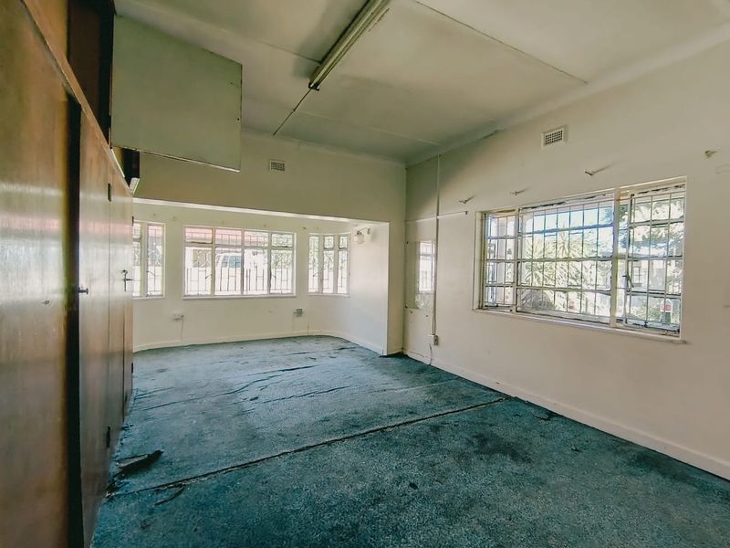 Spacious living area featuring built-in cupboards, large steel-framed windows with security bars, and original flooring in need of renovation.