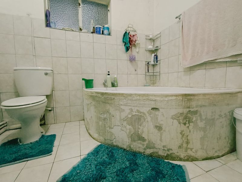 A bathroom featuring a built-in bathtub and toilet with tiled surfaces. The room requires cosmetic updates and repairs to the bathtub surround and wall finishes.
