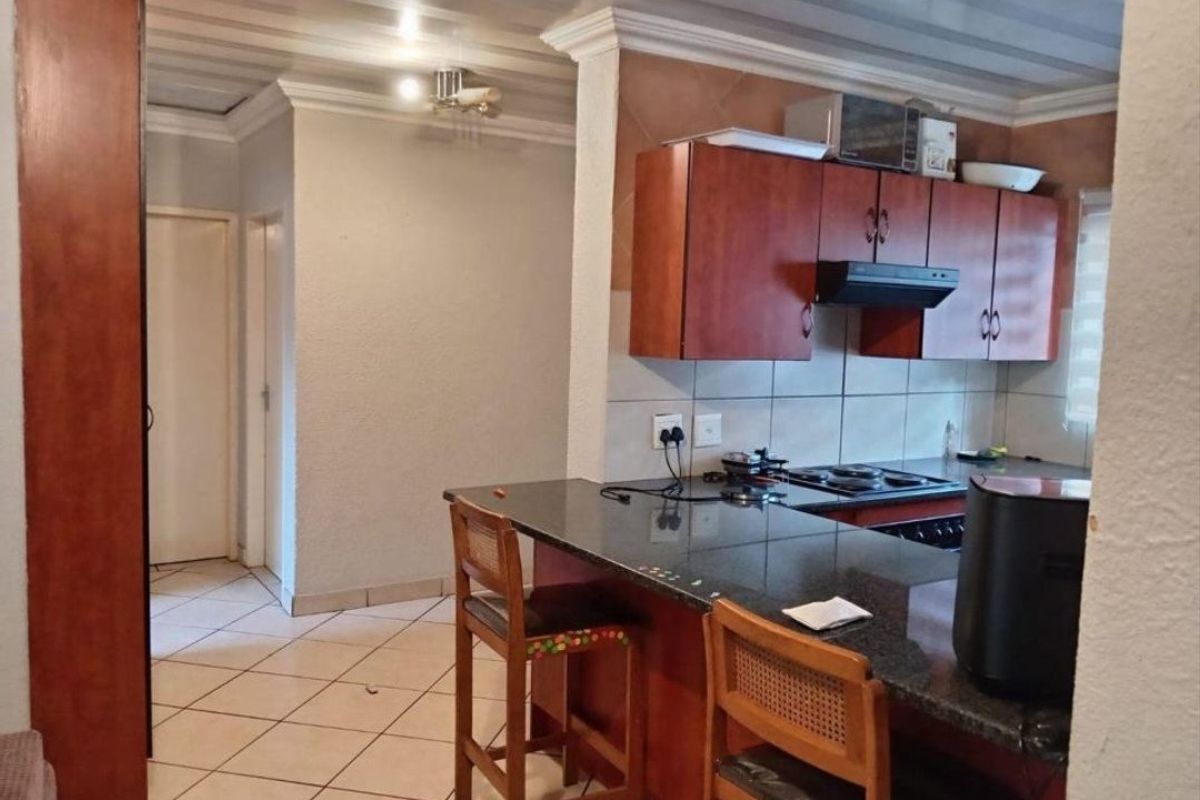 A functional kitchen featuring wooden cabinetry, granite countertops, a built-in hob with extractor fan, and tiled flooring, opening into a hallway area.