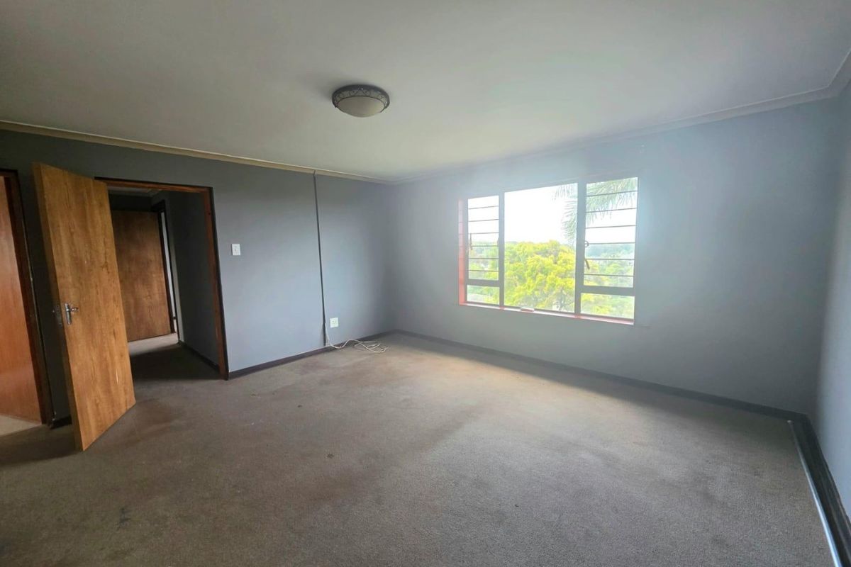 An empty bedroom featuring carpet flooring, a flush mount ceiling light, and louvered windows with a view of the surrounding greenery.