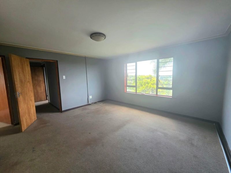 An empty bedroom featuring carpet flooring, a flush mount ceiling light, and louvered windows with a view of the surrounding greenery.