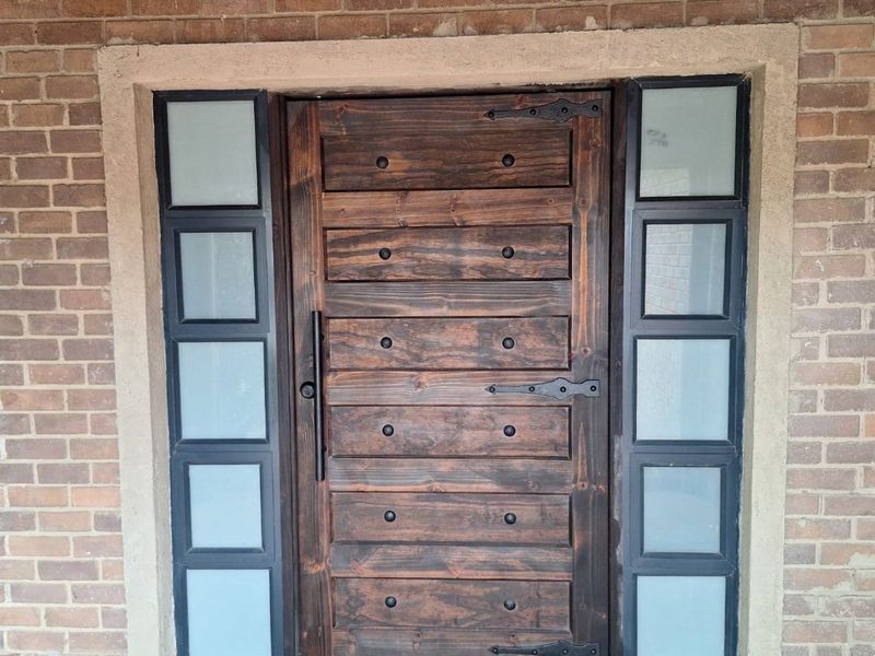 A rustic timber front door featuring decorative iron hinges and vertical pull handle, flanked by modern frosted glass sidelights set into an exposed brick facade.