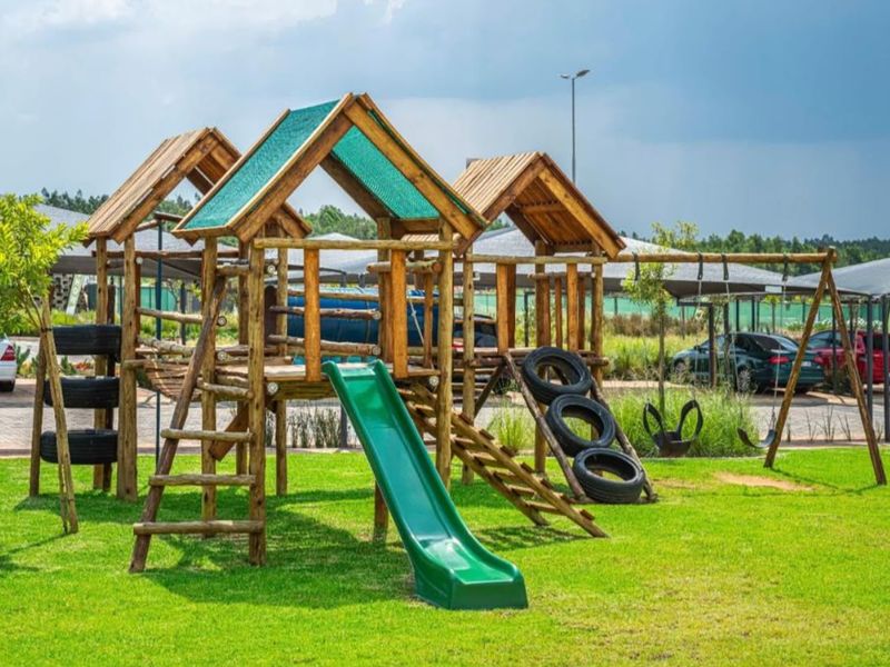 A well-maintained communal garden area featuring a wooden playground structure with a slide and swing set, set against a paved parking area.