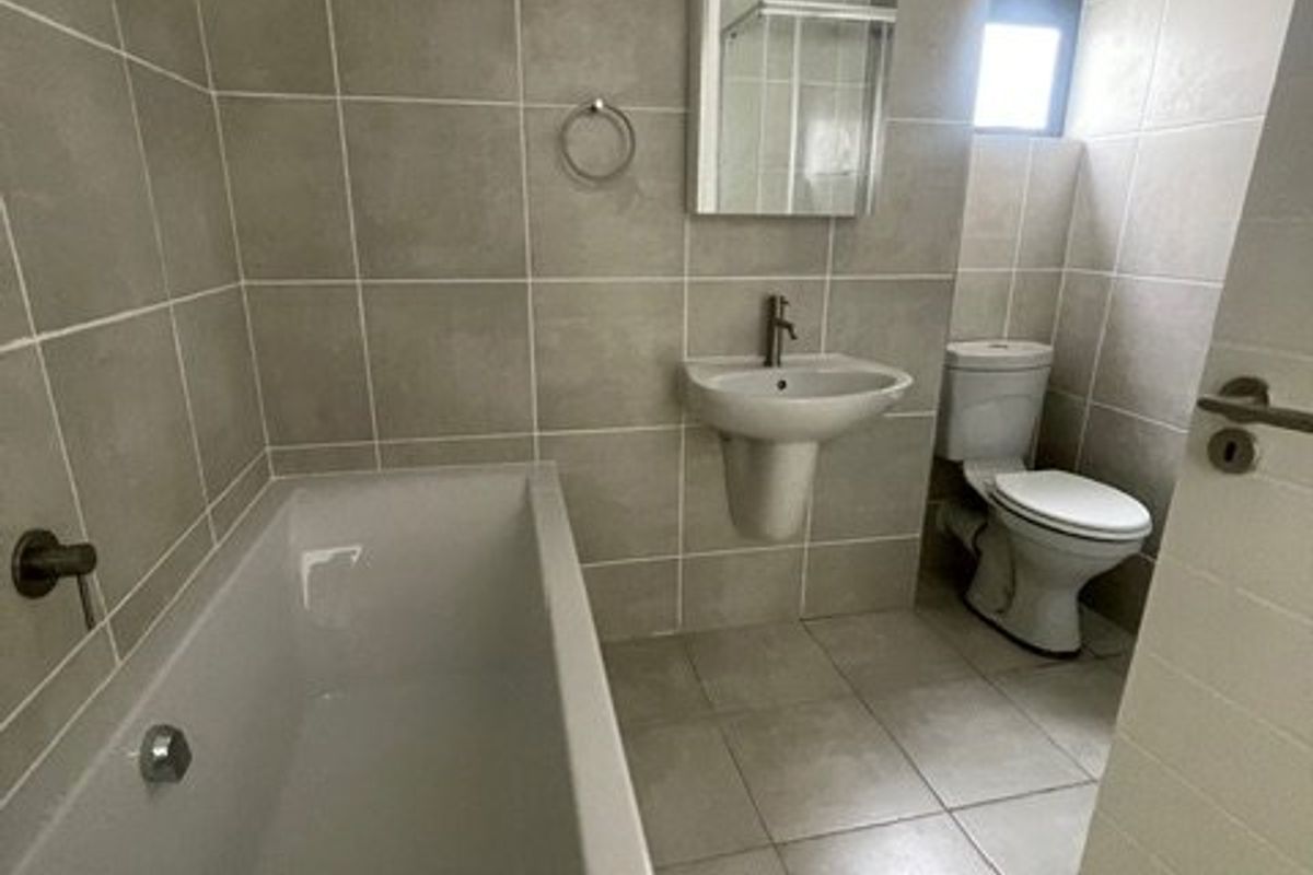 A clean, neutral-toned bathroom featuring a full-size bathtub, pedestal sink, and toilet with modern wall and floor tiling.
