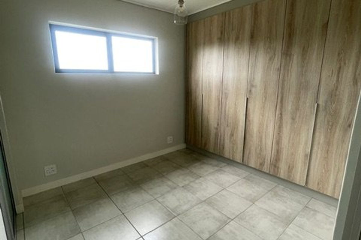 A clean, empty bedroom featuring modern wood-finish built-in cupboards, neutral tiled flooring, and a simple pendant light fixture.