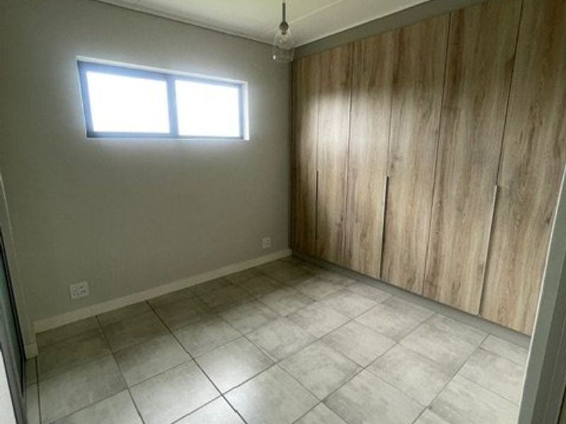 A clean, empty bedroom featuring modern wood-finish built-in cupboards, neutral tiled flooring, and a simple pendant light fixture.