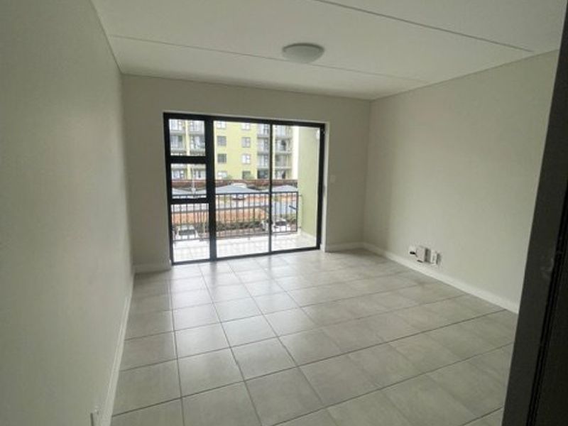A bright, empty living area featuring neutral tiled flooring and a large sliding glass door providing access to a private balcony.