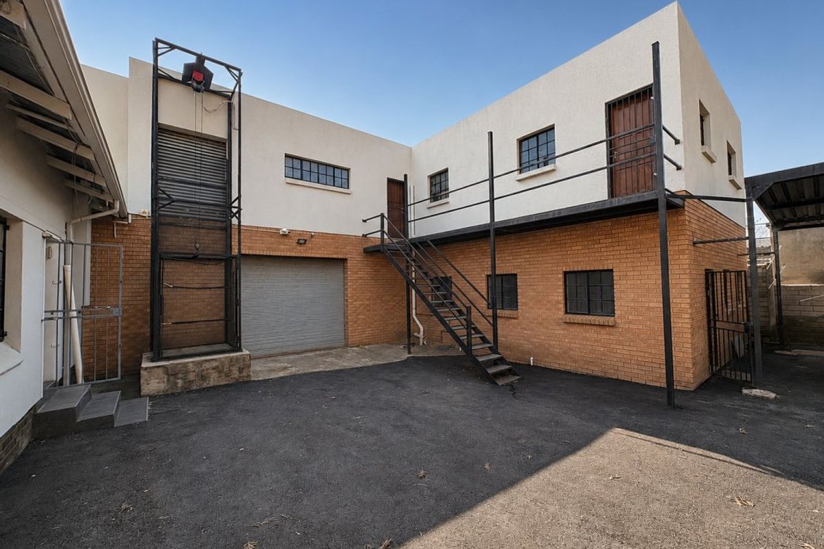 Rear exterior view of a commercial property featuring a paved yard, industrial roller shutter door, external metal staircase, and balcony access.