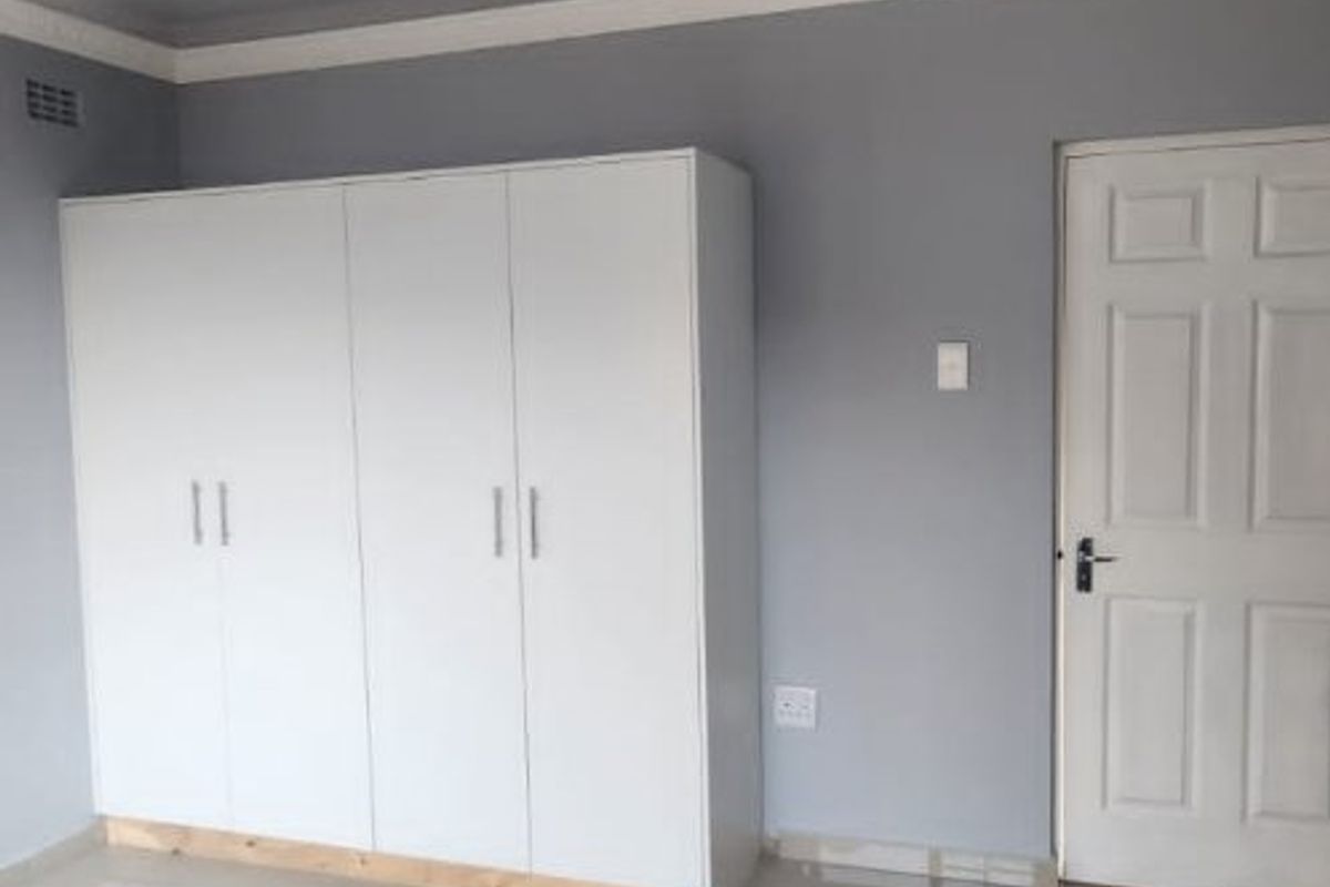 A clean, empty bedroom featuring neutral grey walls, large format floor tiles, a panelled ceiling, and a modern white built-in wardrobe.