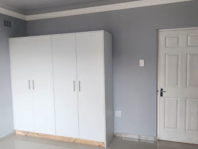 A clean, empty bedroom featuring neutral grey walls, large format floor tiles, a panelled ceiling, and a modern white built-in wardrobe.