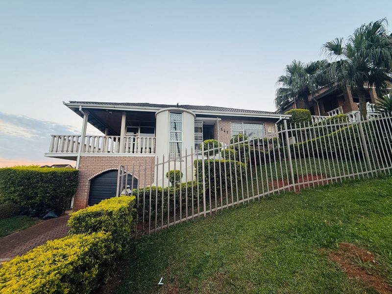 Front elevation of a brick house featuring a raised balcony, secure perimeter fencing, and an integrated garage on a sloped residential lot.
