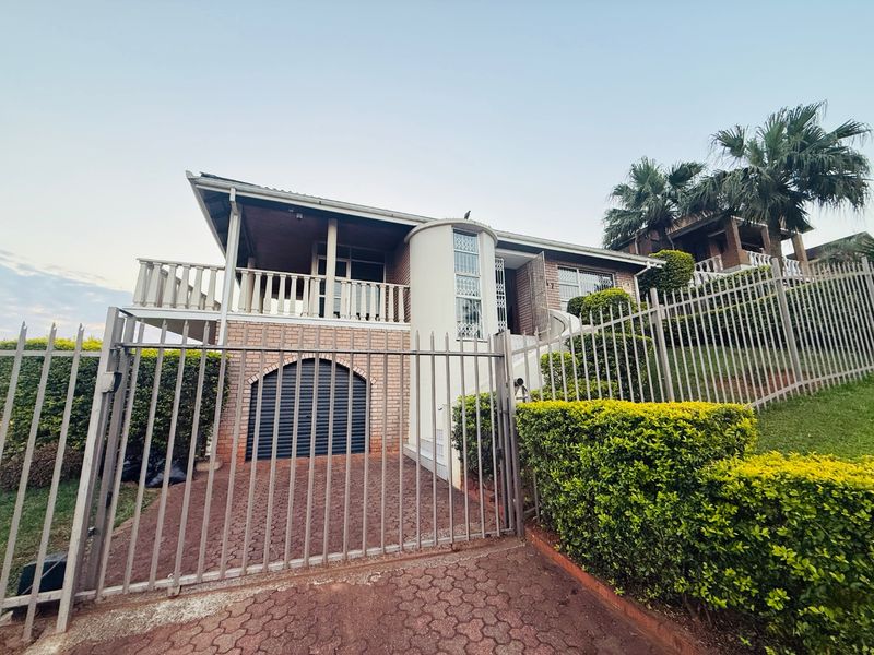 Front view of a multi-level brick house featuring a paved driveway, secure metal fencing, a balcony, and a roller garage door.