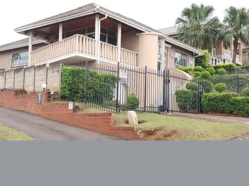 Front elevation of a multi-level brick house featuring a balcony, security fencing, and a paved driveway.