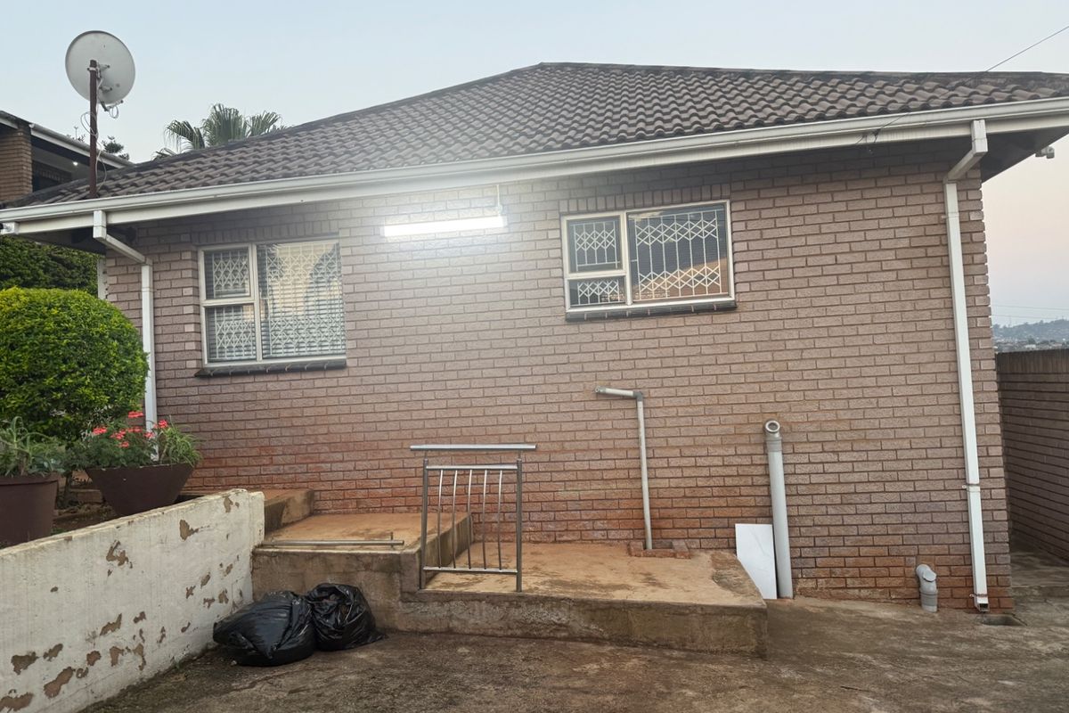 Rear exterior view of the property featuring a brick facade, tiled roof, security-fitted windows, and a concrete patio area with a stainless steel handrail.