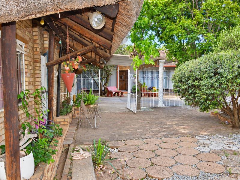 A rustic outdoor patio area featuring a thatched roof, brick exterior walls, and a paved courtyard with established garden landscaping.