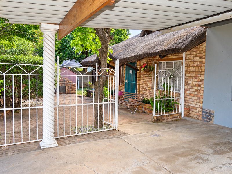A covered patio area featuring a thatched roof, brick facade, and decorative column, overlooking a paved garden space with a built-in brick braai.